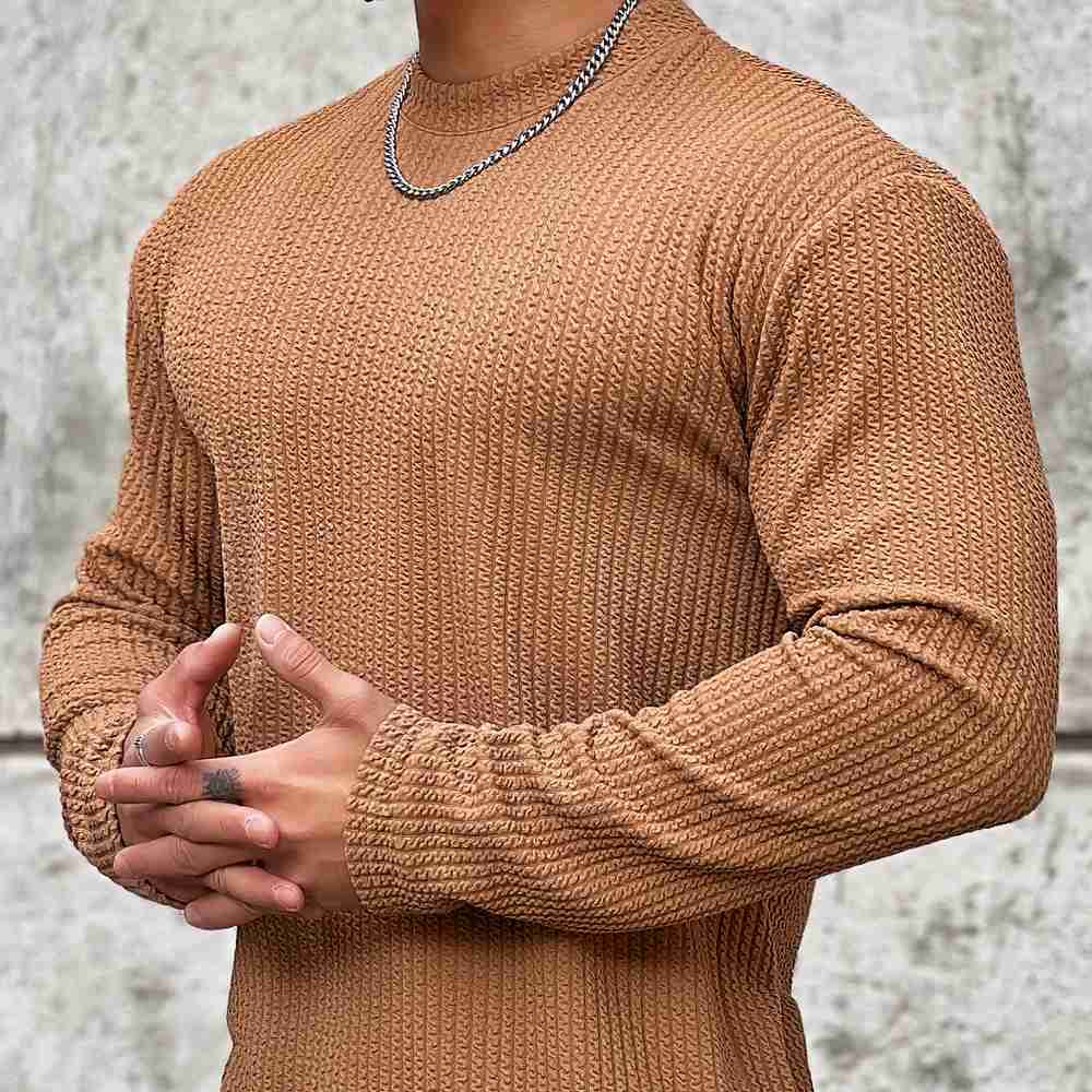 Homme portant un pull marron texturé, chaîne argentée visible, posant devant un mur gris. Mode masculine, style automne-hiver, tendance actuelle.