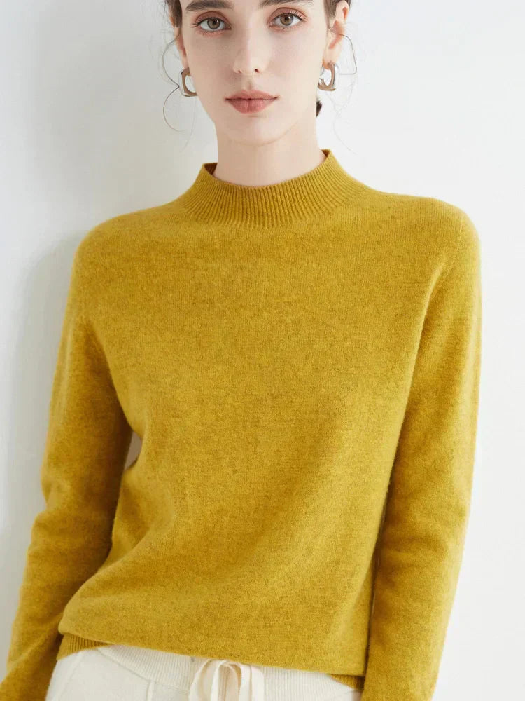 Femme portant un pull en cachemire jaune moutarde, manches longues, col montant, posant contre un mur blanc. Mode automne-hiver élégante et confortable.