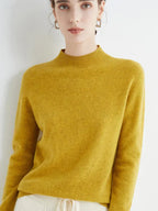 Femme portant un pull en cachemire jaune moutarde, manches longues, col montant, posant contre un mur blanc. Mode automne-hiver élégante et confortable.