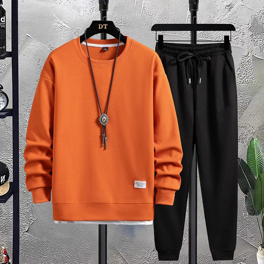 Sweat-shirt orange et pantalon noir sur cintres, style décontracté, fond gris texturé, collier pendentif, mode homme automne-hiver.