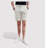 Homme portant un short beige élégant, ceinture assortie, t-shirt noir et baskets blanches, posant sur fond blanc. Mode masculine estivale.
