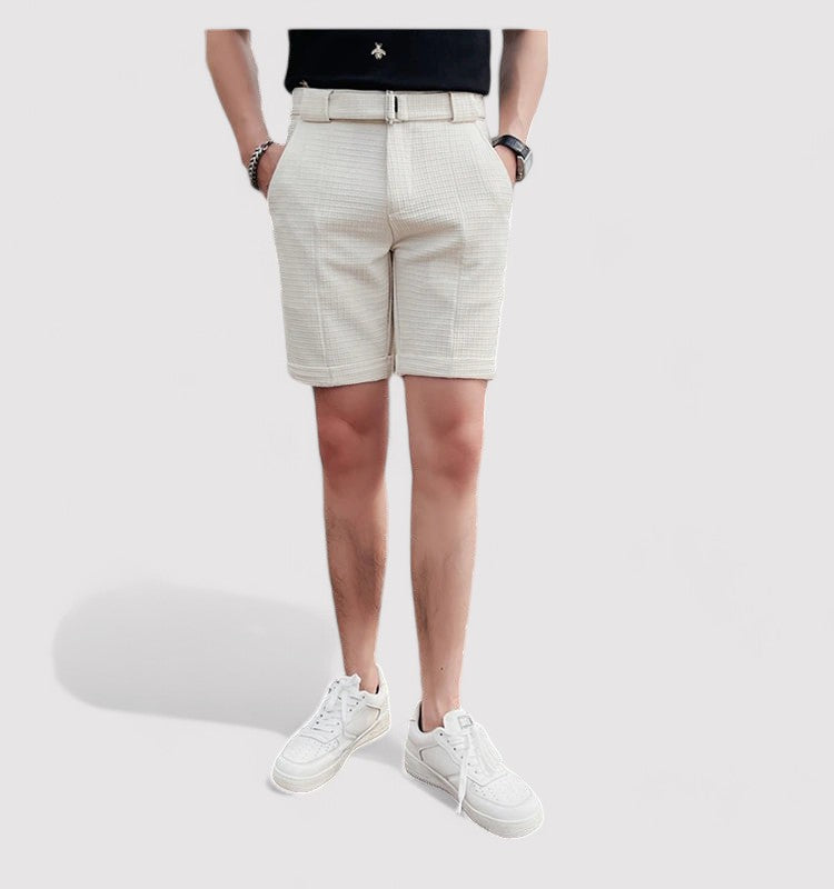 Homme portant un short beige élégant, ceinture assortie, t-shirt noir et baskets blanches, posant sur fond blanc. Mode masculine estivale.