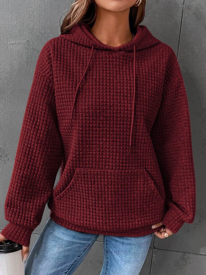 Femme portant un sweat à capuche rouge bordeaux texturé, manches longues, avec poche kangourou, tenue décontractée, mur gris en arrière-plan.