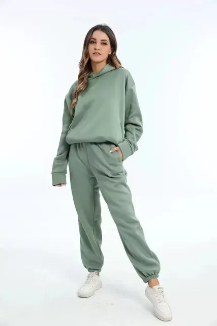 Femme portant un ensemble de survêtement vert, composé d'un sweat à capuche et d'un pantalon, avec des baskets blanches. Mode décontractée tendance.