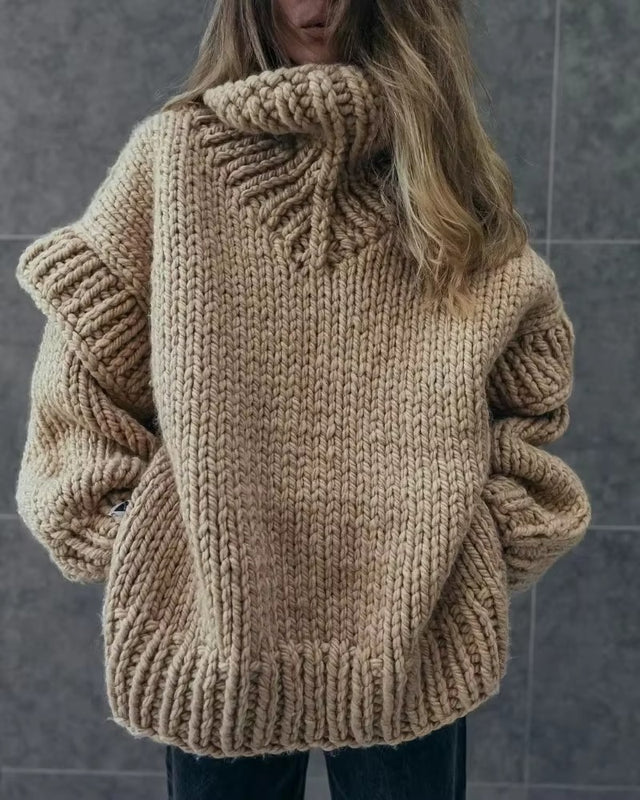 Femme portant un pull en laine épais beige, tricoté à la main, style oversize, devant un mur gris. Mode hiver, confort, tendance.