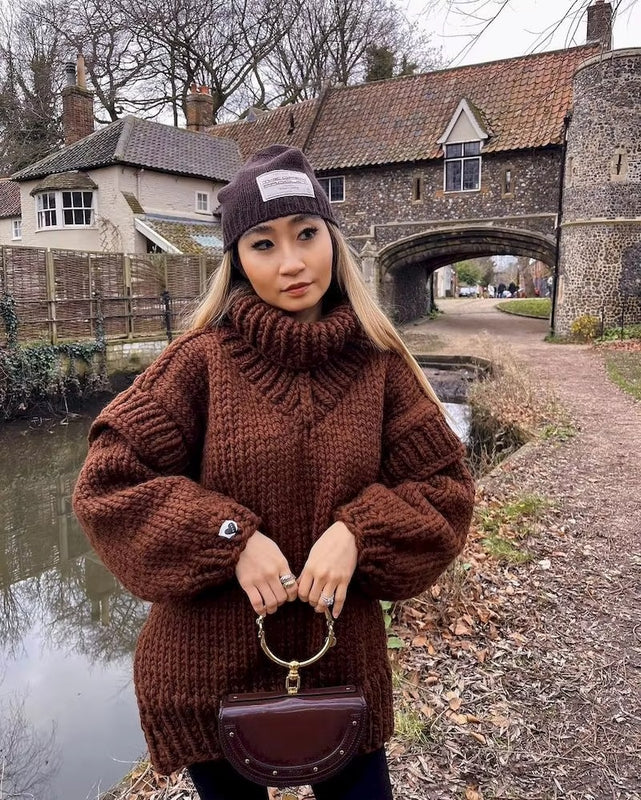 Femme portant un pull en laine marron et bonnet, posant devant un pont en pierre et une maison ancienne, style automnal, mode hiver.