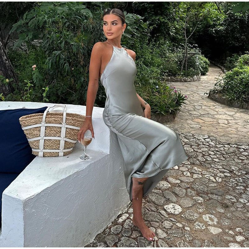 Femme en robe satinée grise, assise sur un muret blanc avec un verre de vin, entourée de verdure. Mode élégante, style estival, détente.