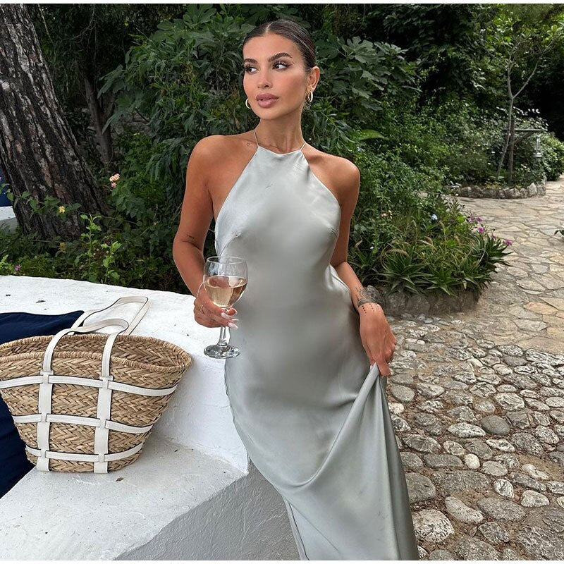 Femme élégante en robe satinée grise tenant un verre de vin, posant dans un jardin luxuriant. Mode chic, soirée d'été, style glamour.