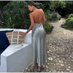 Femme en robe satinée argentée, dos nu, debout sur une terrasse en pierre, près d'un sac en osier et d'un verre de vin, entourée de verdure luxuriante.