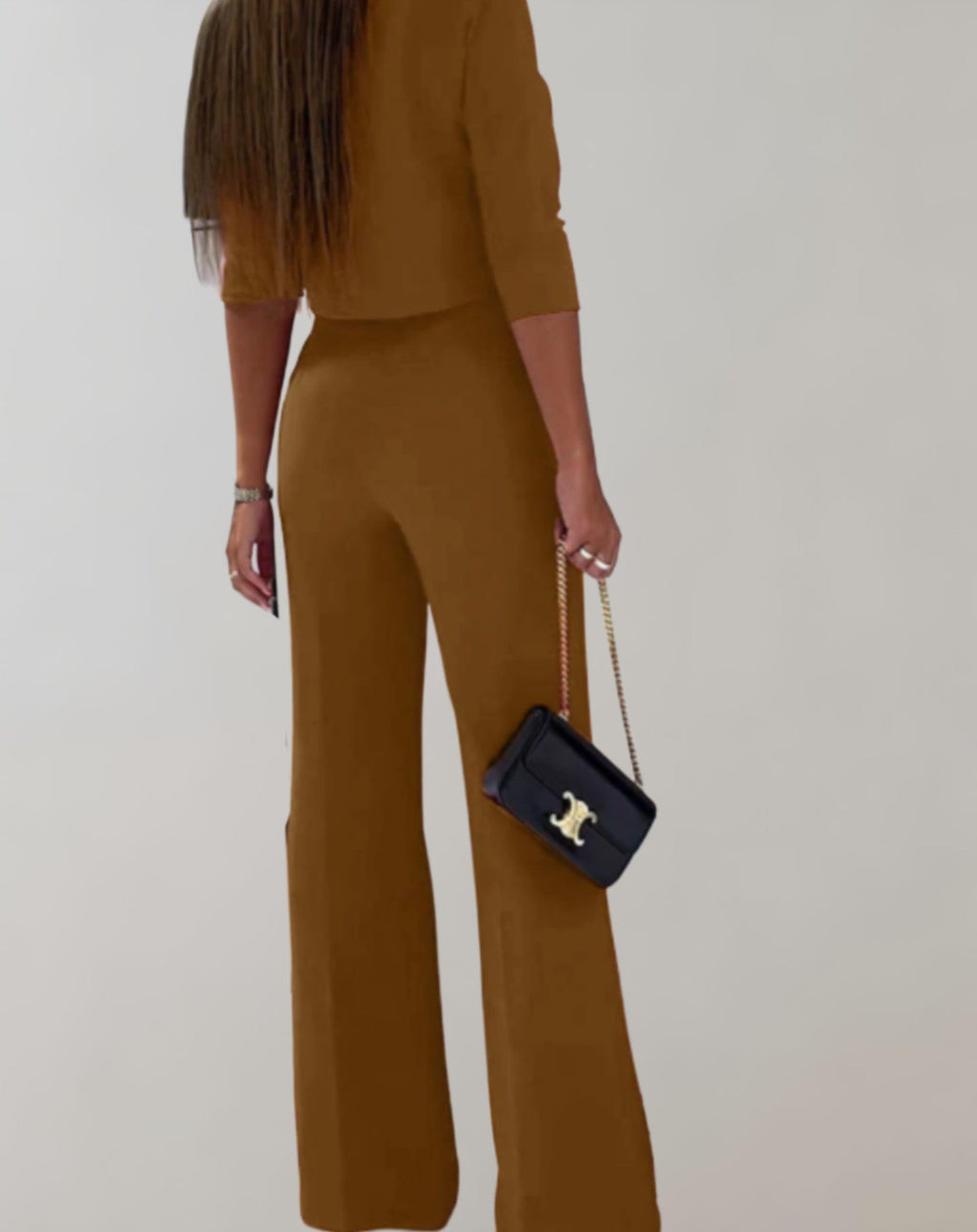 Femme en tenue marron élégante, portant un sac à main noir avec chaîne dorée, cheveux longs lisses, posant sur fond blanc minimaliste.