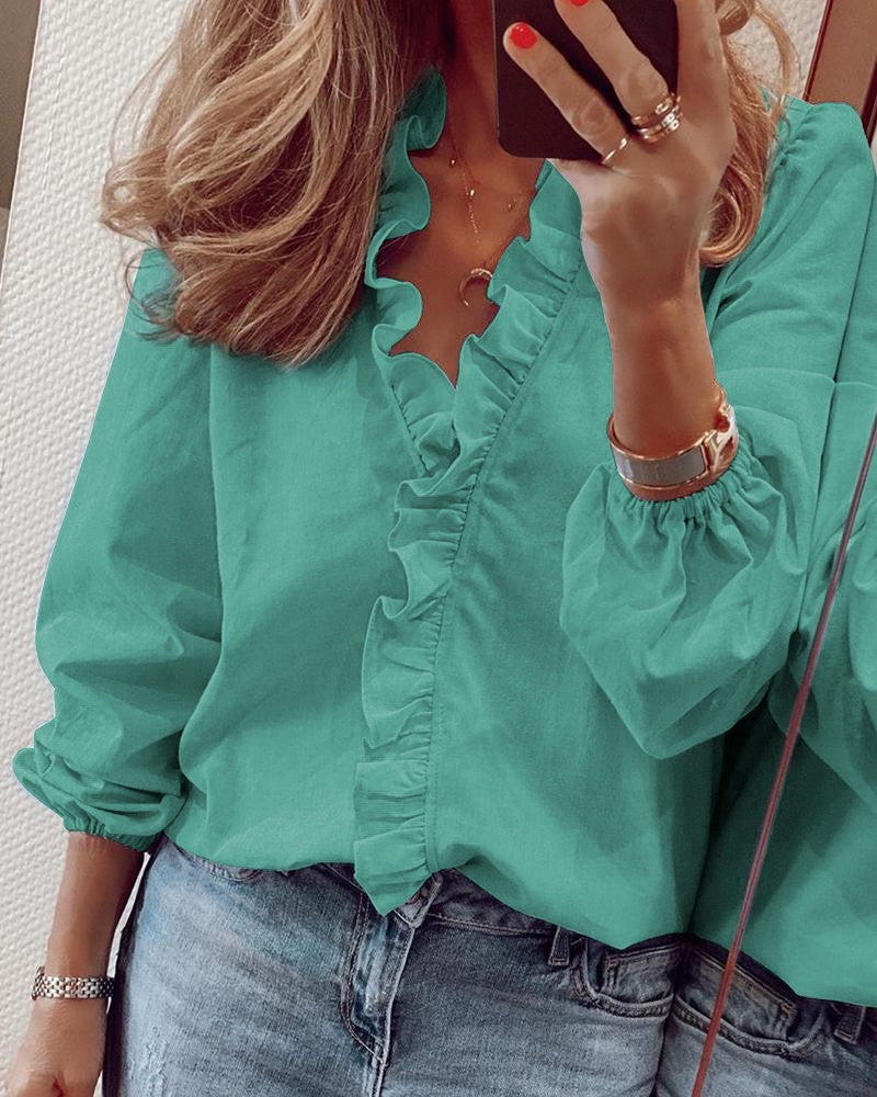 Femme portant une blouse verte à volants, manches longues, avec un collier lune et montre dorée, tenant un smartphone, reflet dans le miroir.