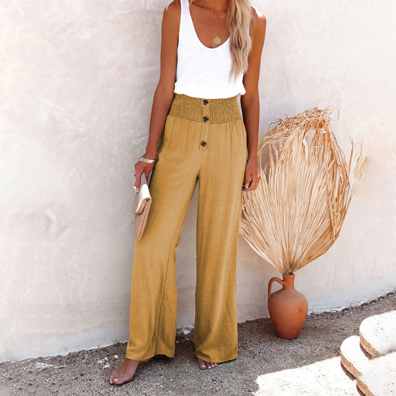 Femme en pantalon large beige taille haute, top blanc, tenant une pochette, posant devant un mur texturé avec vase et plante séchée. Mode estivale chic.