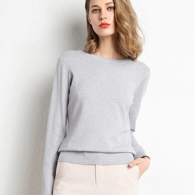 Femme portant un pull gris élégant, manches longues, col rond, posant contre un mur blanc. Mode féminine, style décontracté chic.