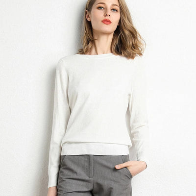 Femme portant un pull blanc élégant et un pantalon gris, posant contre un mur blanc. Mode féminine chic, tenue décontractée moderne.