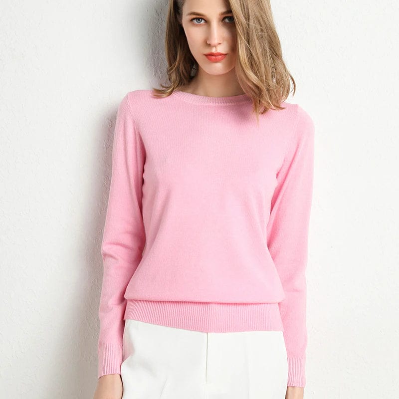 Jeune femme portant un pull rose en cachemire, posant contre un mur blanc. Mode féminine élégante, tenue automne-hiver, style décontracté chic.