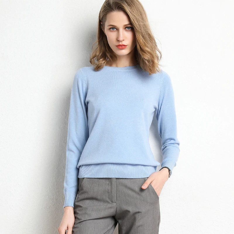 Femme portant un pull bleu clair et un pantalon gris, posant contre un mur blanc. Mode féminine élégante, tenue décontractée chic.