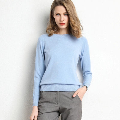 Femme portant un pull bleu clair et un pantalon gris, posant contre un mur blanc. Mode féminine élégante, tenue décontractée chic.