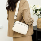 Femme en tailleur beige portant un sac à bandoulière blanc avec sangle à motifs, fleurs blanches en arrière-plan. Mode élégante et moderne.