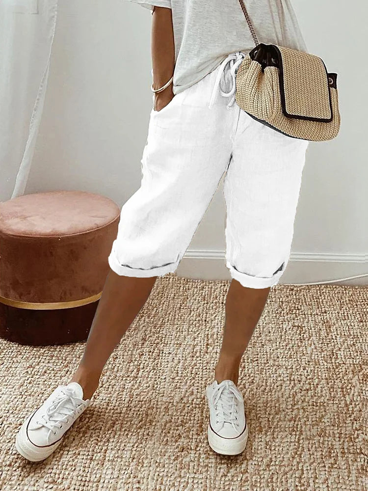 Femme portant un short blanc décontracté, t-shirt gris, baskets blanches et sac à main en paille, posant sur un tapis beige. Mode estivale chic.