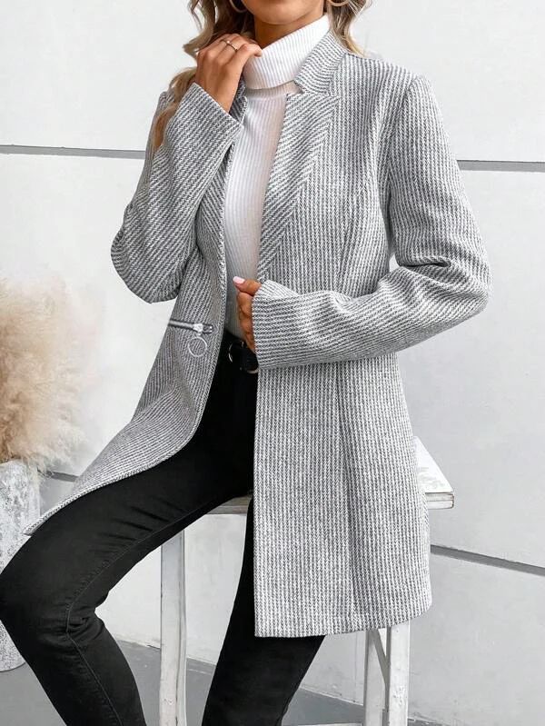 Femme portant un blazer gris à rayures, col roulé blanc et pantalon noir, posant contre un mur moderne. Mode élégante et tendance automne-hiver.