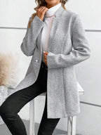 Femme portant un blazer gris à rayures, col roulé blanc et pantalon noir, posant contre un mur moderne. Mode élégante et tendance automne-hiver.