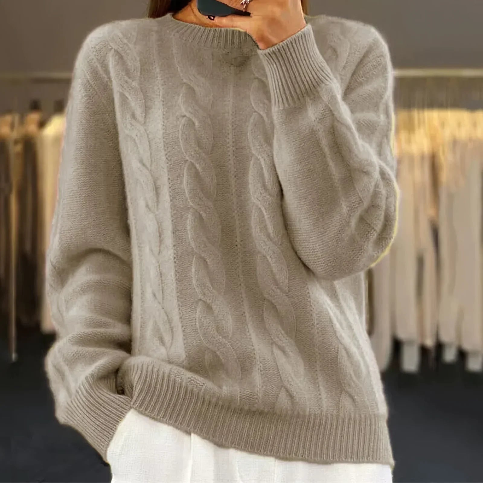 Femme portant un pull en laine torsadé beige, manches longues, col rond, devant un fond flou de vêtements suspendus. Mode automne-hiver élégante.