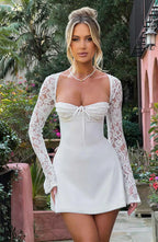 Femme en robe blanche élégante avec manches en dentelle, posant dans un jardin luxuriant. Mode féminine, style chic, tenue de soirée.
