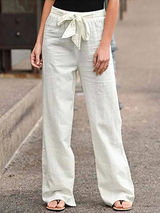 Pantalon large en lin blanc avec ceinture nouée, porté par une femme en sandales noires, sur un trottoir urbain. Mode décontractée estivale.