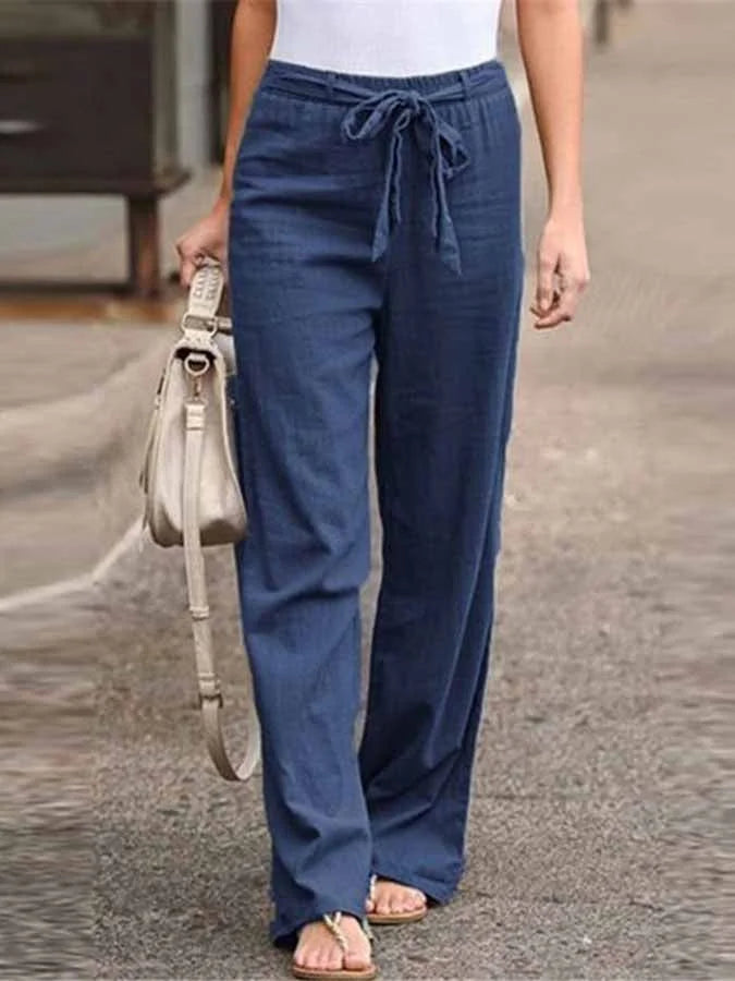 Pantalon large bleu en lin pour femme, avec ceinture à nouer, porté avec un haut blanc et des sandales, sac à main beige en bandoulière.