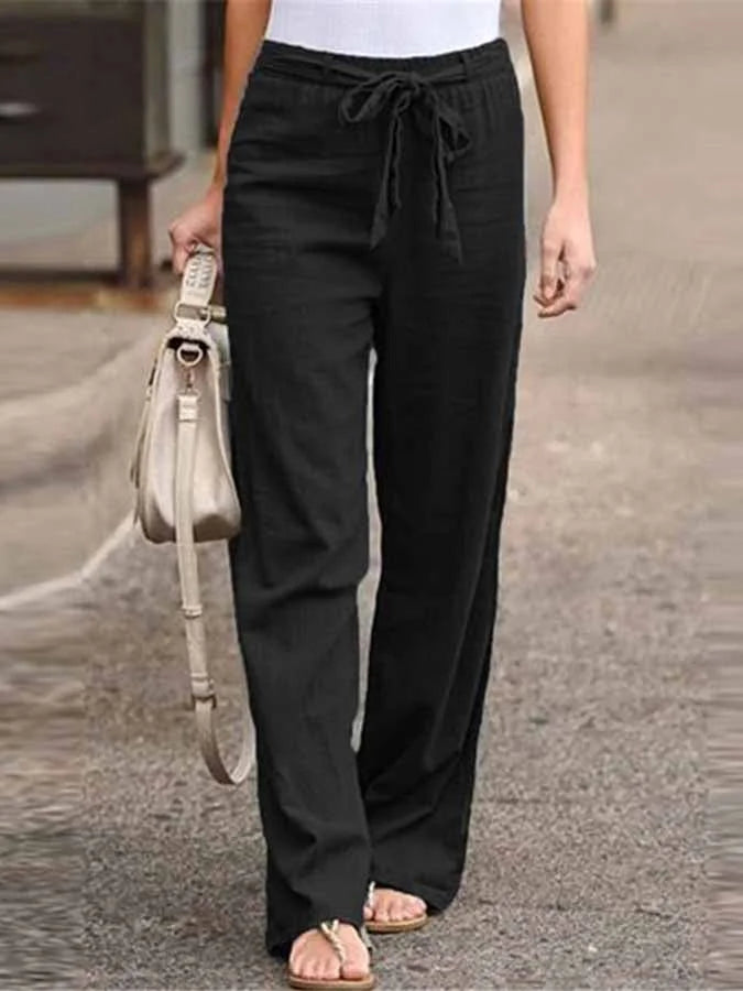 Pantalon noir ample pour femme avec ceinture nouée, porté avec un haut blanc et des sandales, accompagné d'un sac à main beige.
