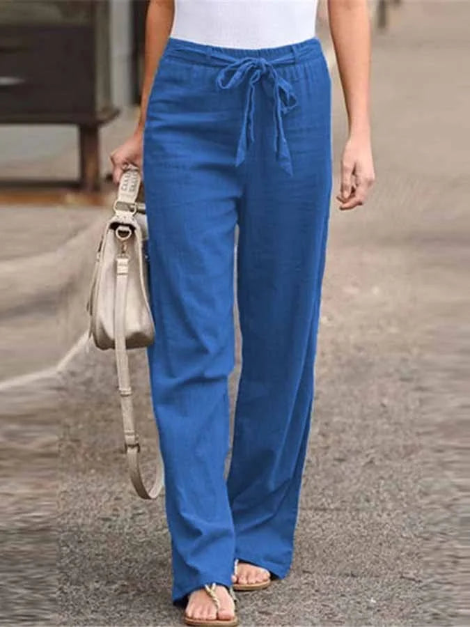 Pantalon bleu ample en lin pour femme, avec ceinture à nouer, porté avec un débardeur blanc et des sandales, sac à main beige en cuir.
