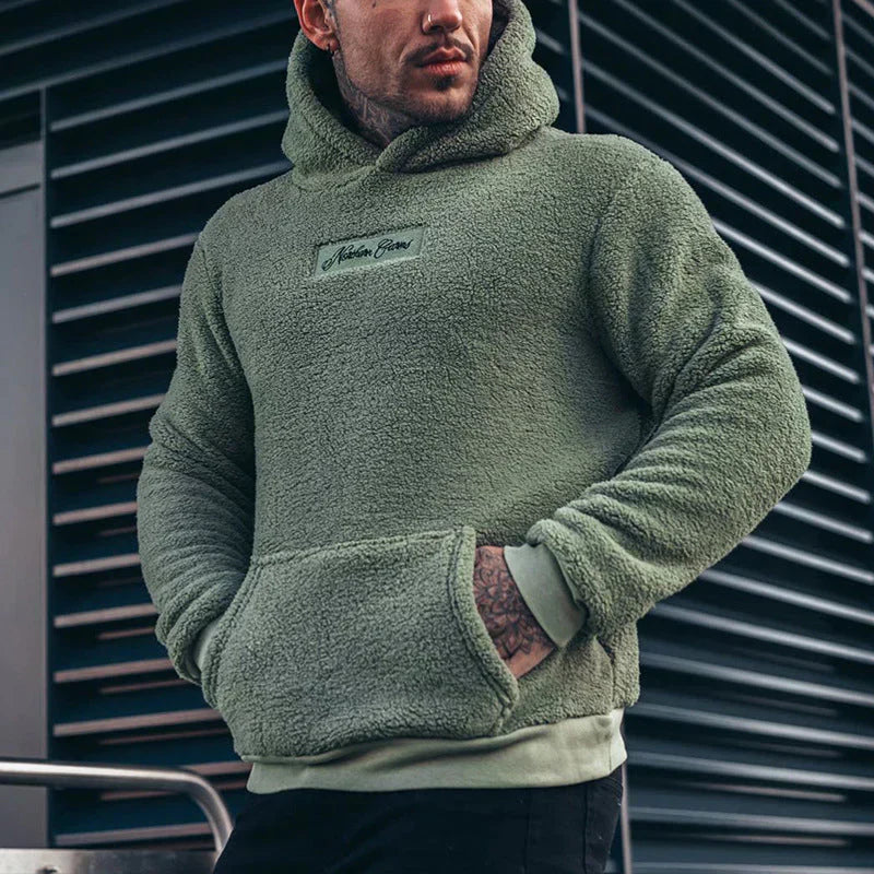 Homme portant un sweat à capuche vert en polaire, devant un mur moderne. Mode urbaine, vêtement chaud, style décontracté.