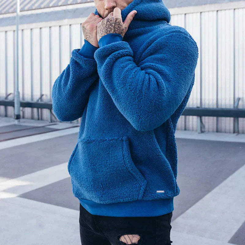 Homme portant un sweat à capuche bleu en polaire, debout à l'extérieur. Mode urbaine, vêtements d'hiver, style décontracté.