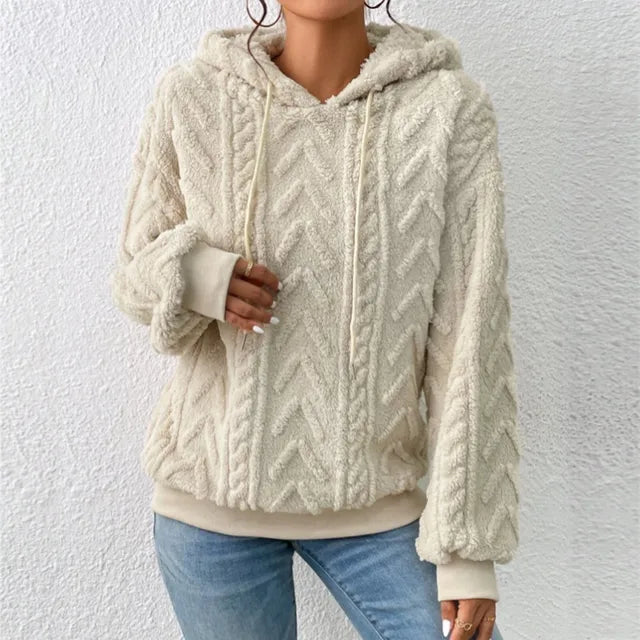 Femme portant un pull à capuche en laine beige texturé, motif chevrons, manches longues, avec jean bleu. Mode automne-hiver, style décontracté.