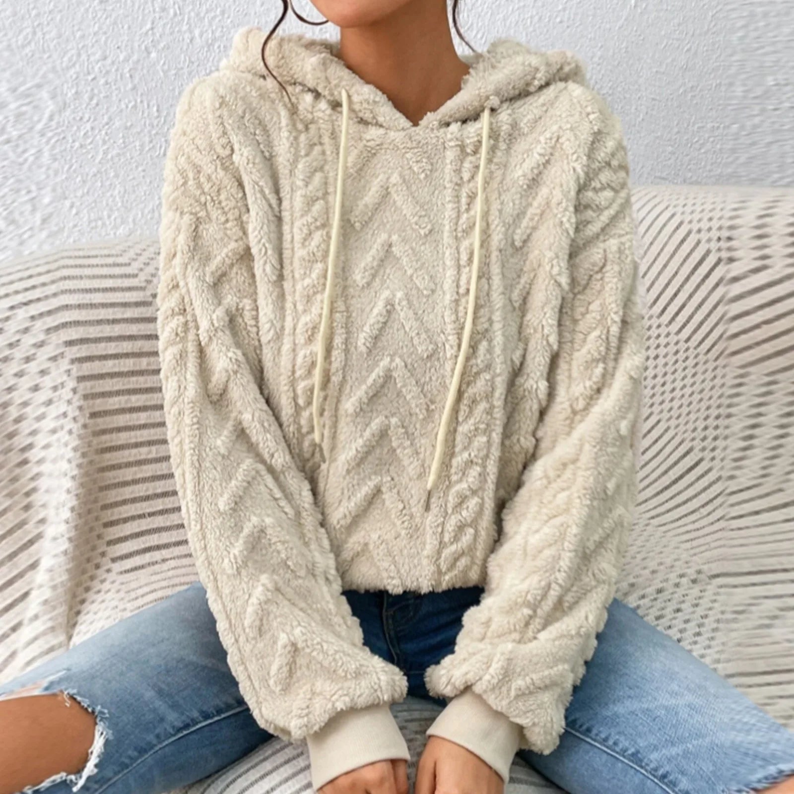 Femme portant un pull à capuche en laine beige texturé, motif chevrons, assise sur un canapé. Mode automne-hiver, confort et style décontracté.
