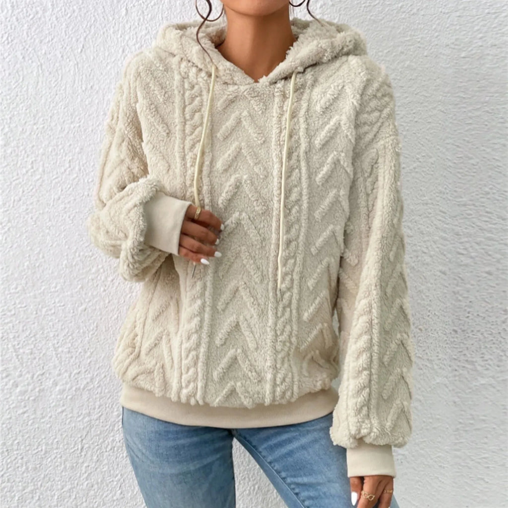 Femme portant un pull à capuche en laine beige texturée, motif chevrons, manches longues, associé à un jean bleu. Mode automne-hiver tendance.