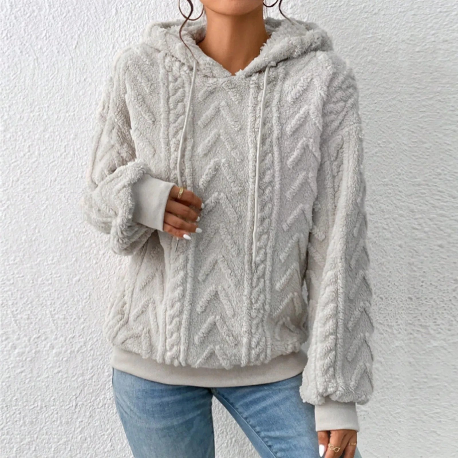 Femme portant un pull à capuche en laine beige texturée, motif torsadé, manches longues, sur fond blanc. Mode automne-hiver, style décontracté.