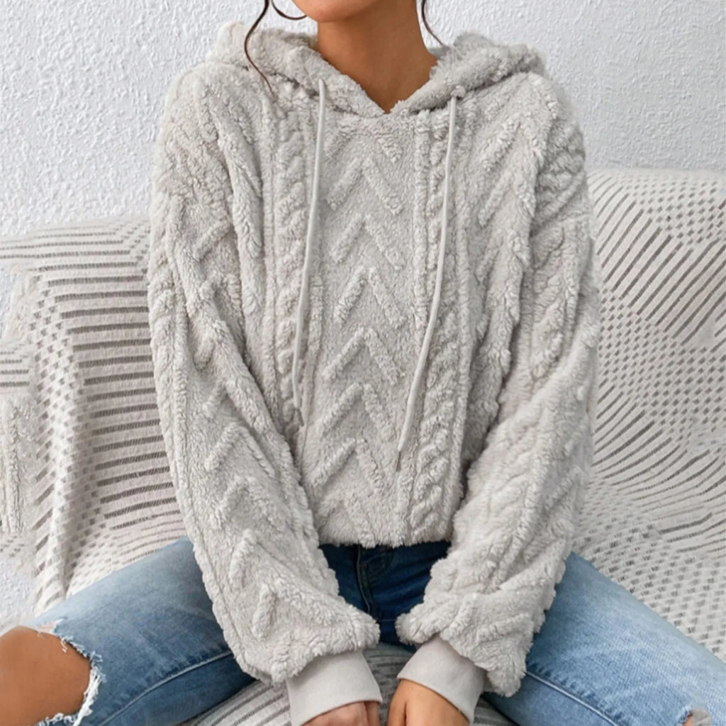 Femme portant un pull à capuche en laine grise texturée, motif chevrons, assise sur un canapé, jeans décontractés. Mode automne-hiver tendance.