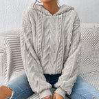 Femme portant un pull à capuche en laine grise texturée, motif chevrons, assise sur un canapé, jeans décontractés. Mode automne-hiver tendance.