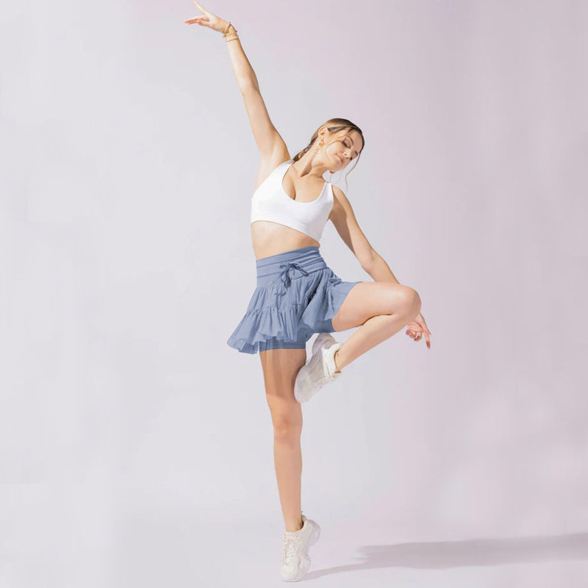 Femme en tenue de sport, brassière blanche et jupe bleue, effectuant une pose de danse sur fond clair. Fitness, danse, mode active.