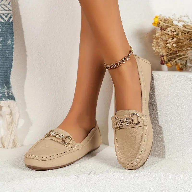 Chaussures mocassins beiges élégantes pour femme, ornées de boucles dorées, posées sur un fond blanc avec un bracelet de cheville doré.