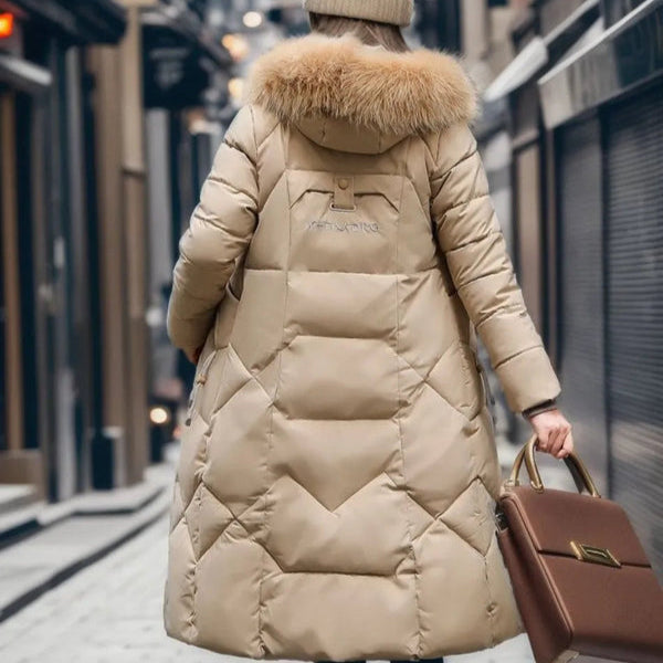 Doudoune longue beige avec capuche en fourrure, femme de dos dans une rue enneigée, tenant un sac à main marron. Mode hiver, manteau chaud.
