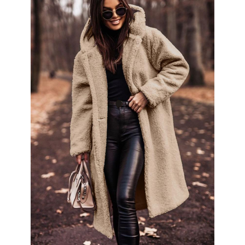 Femme portant un manteau en fausse fourrure beige, lunettes de soleil, pantalon en cuir noir, tenant un sac à main, marchant dans un parc automnal.