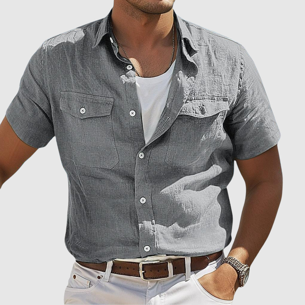 Homme portant une chemise grise à manches courtes, avec un t-shirt blanc en dessous, ceinture marron et pantalon blanc. Mode masculine décontractée.