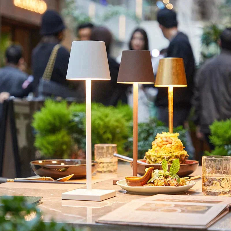 Trois lampes de table modernes éclairent un repas sur une table de restaurant, avec des plats appétissants et des boissons, en arrière-plan flou de clients.