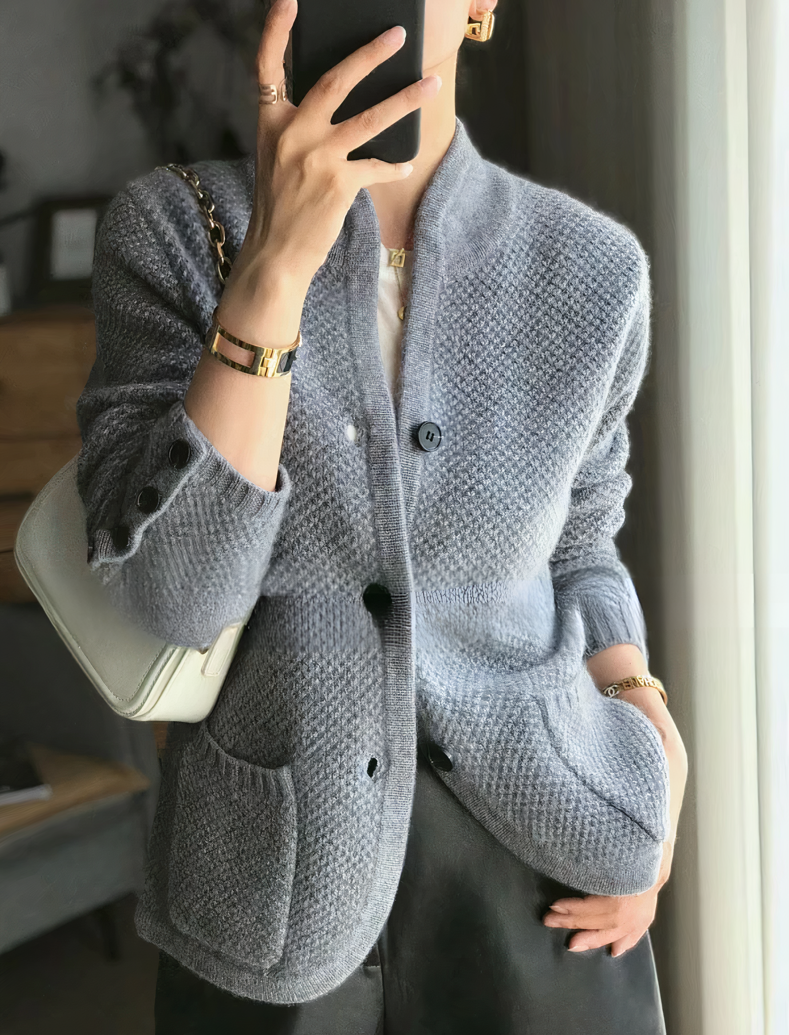Femme portant un cardigan gris texturé, sac blanc à l'épaule, bracelets dorés, tenant un smartphone. Mode élégante, tenue automne-hiver.