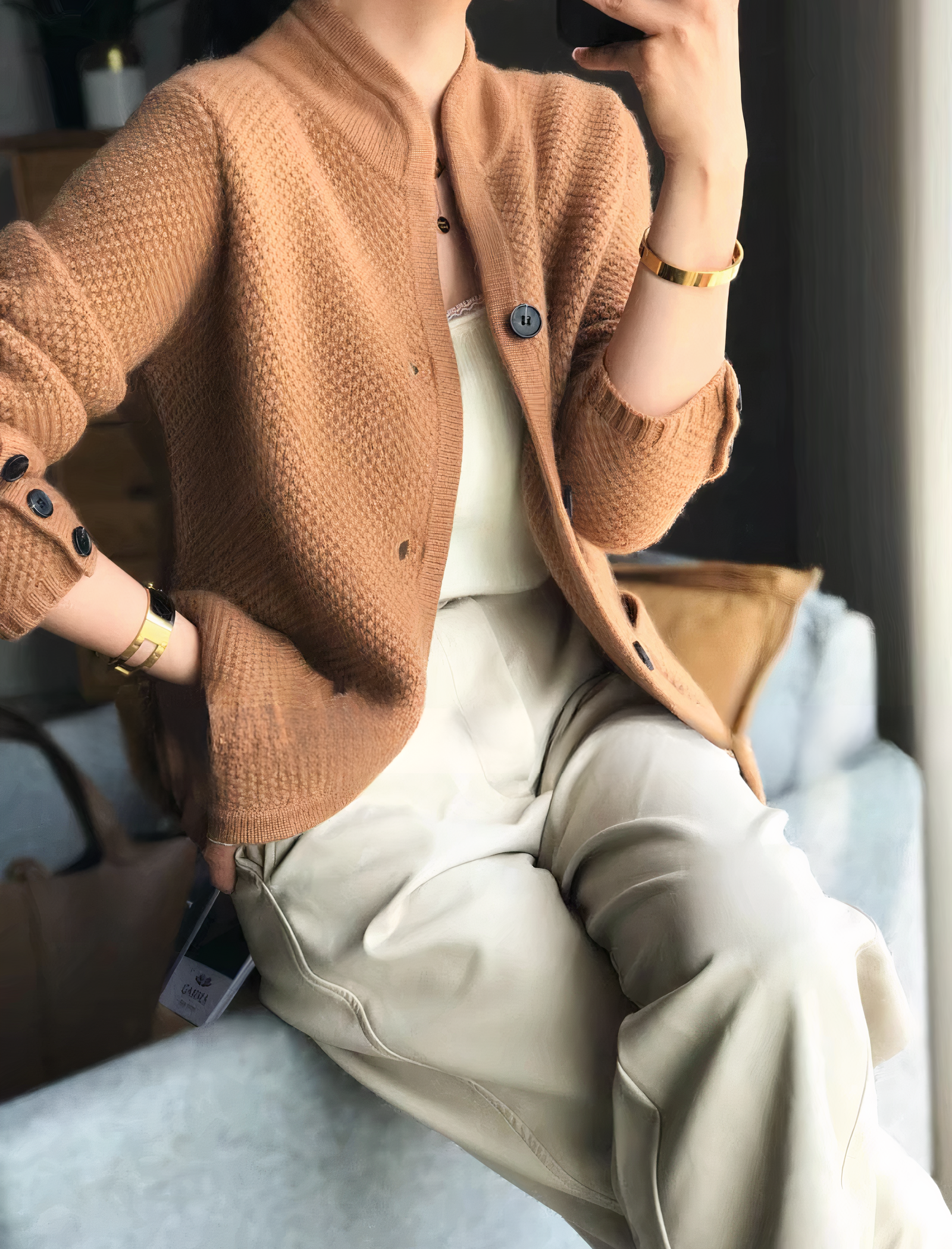 Femme portant un cardigan en laine marron, chemise blanche et pantalon beige, assise sur un canapé. Mode automne, style décontracté chic.