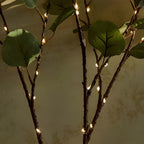 Branche décorative avec feuilles vertes et guirlande lumineuse LED, éclairage d'ambiance intérieur, décoration naturelle et moderne.