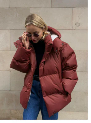Femme portant une doudoune rouge épaisse, lunettes de soleil, et jeans bleus devant un mur beige. Mode hiver, style urbain, tendance vêtements chauds.