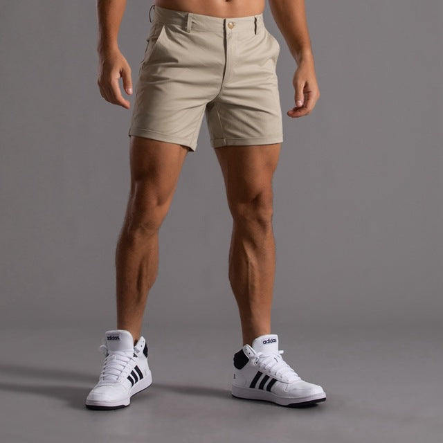 Homme portant un short beige et des baskets blanches Adidas sur fond gris. Mode masculine, tenue décontractée, style sportif.